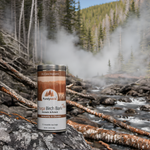 A canister of Purely Wild Chaga Birch Bark Tea displayed in a serene forest, surrounded by birch trees and raw chaga, representing its natural and wild origin as inspired by Dr. Cass Ingram.