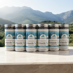 Six tins of PurelyWILD One Needle Tea on a wooden surface with a mountainous background.